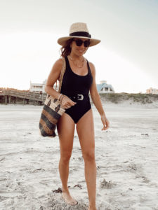 one piece belted black swimsuit with patterned jute tote beach bag and straw hat